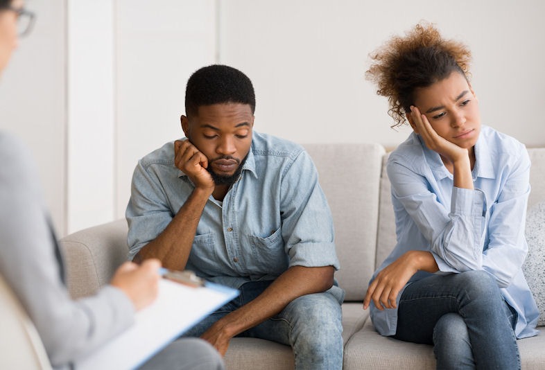 Heterosexual couple wearing similar jeans outfits sitting on a white couch during couples counseling