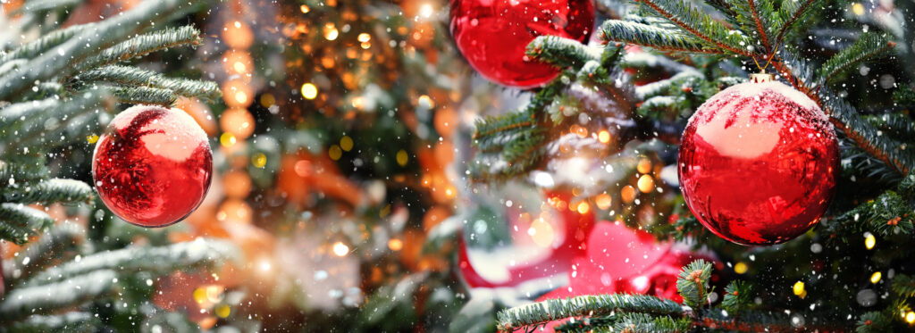 Wide view of pine trees decorated with red ball Christmas ornaments in front of hazy view of decorated storefront as snow falls