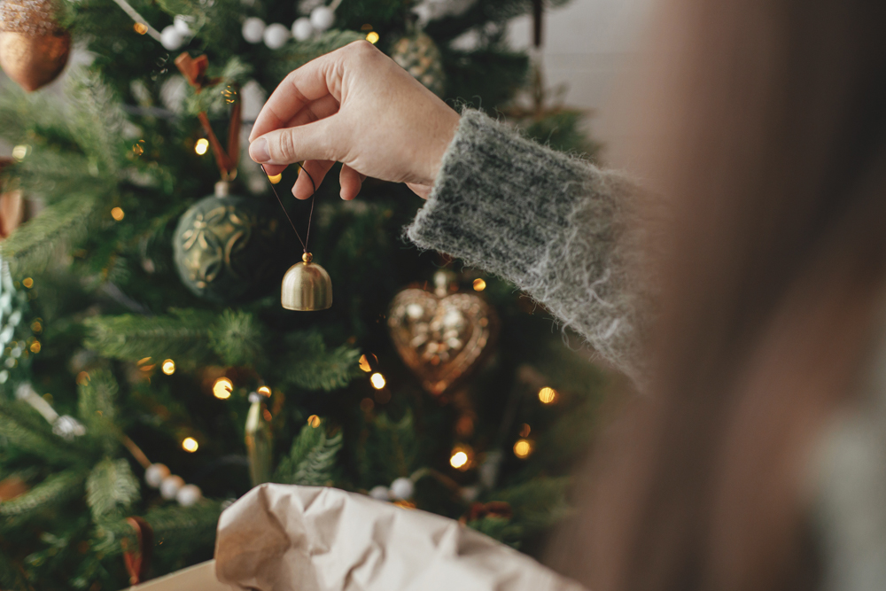 Over-the-shoulder view of a woman wearing a grey sweater decorating a Christmas tree with ornaments