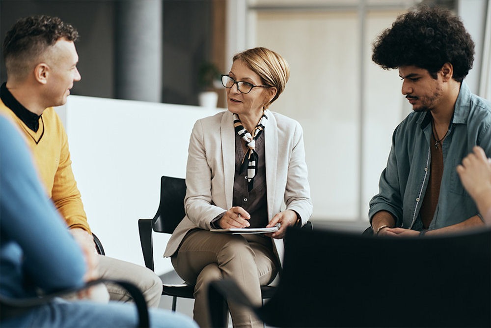 Therapist with glasses taking notes while men talk in a bipolar support group
